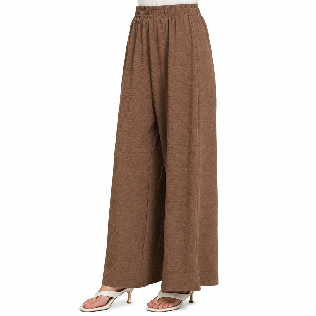 A pair of wide-leg, high-waisted pants in a textured brown fabric, shown on a model from the waist down wearing white heeled sandals against a plain white background.