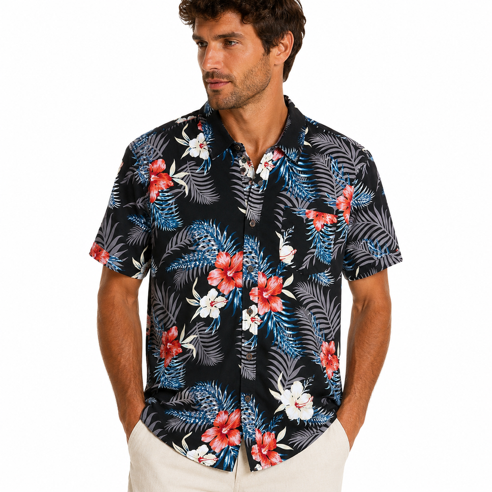 A man wearing a short-sleeve button-up Hawaiian shirt with a tropical floral and palm leaf pattern in red, white, and blue on a black background, paired with off-white pants, against a plain white studio background.