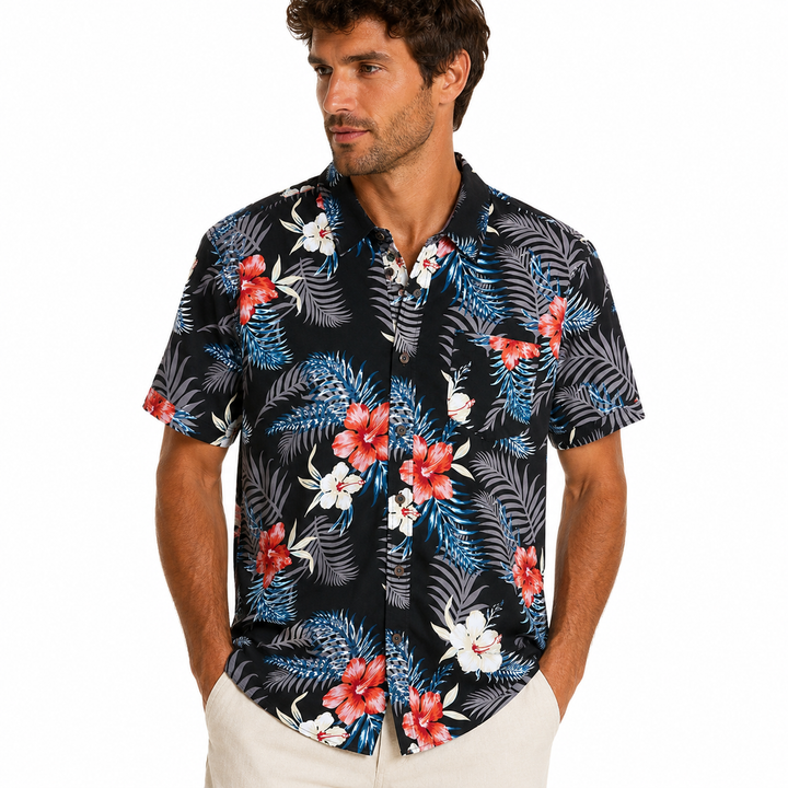 A man wearing a short-sleeve button-up Hawaiian shirt with a tropical floral and palm leaf pattern in red, white, and blue on a black background, paired with off-white pants, against a plain white studio background.