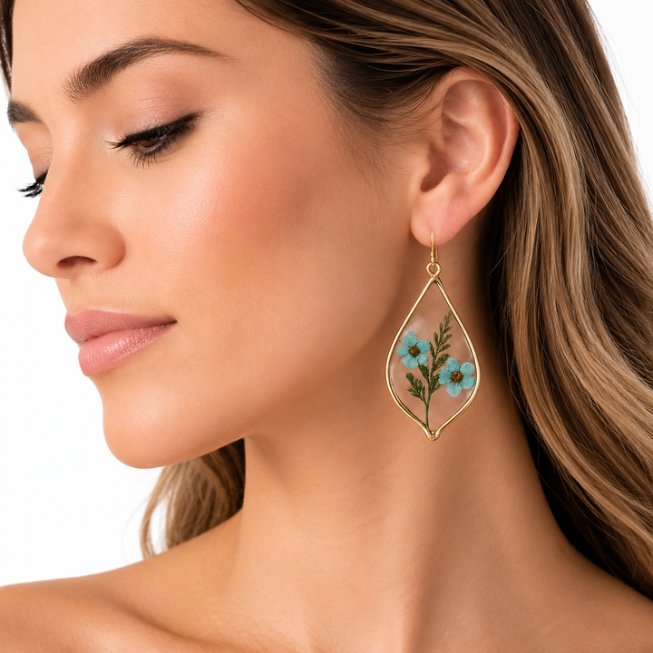 A close-up of a woman wearing a gold teardrop-shaped dangle earring with a preserved blue flower and green leaves inside.