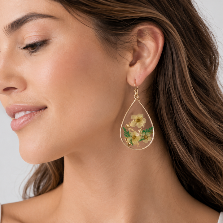 A close-up of a woman wearing a gold teardrop-shaped dangle earring with a clear resin frame containing small yellow and white flowers with green leaves.