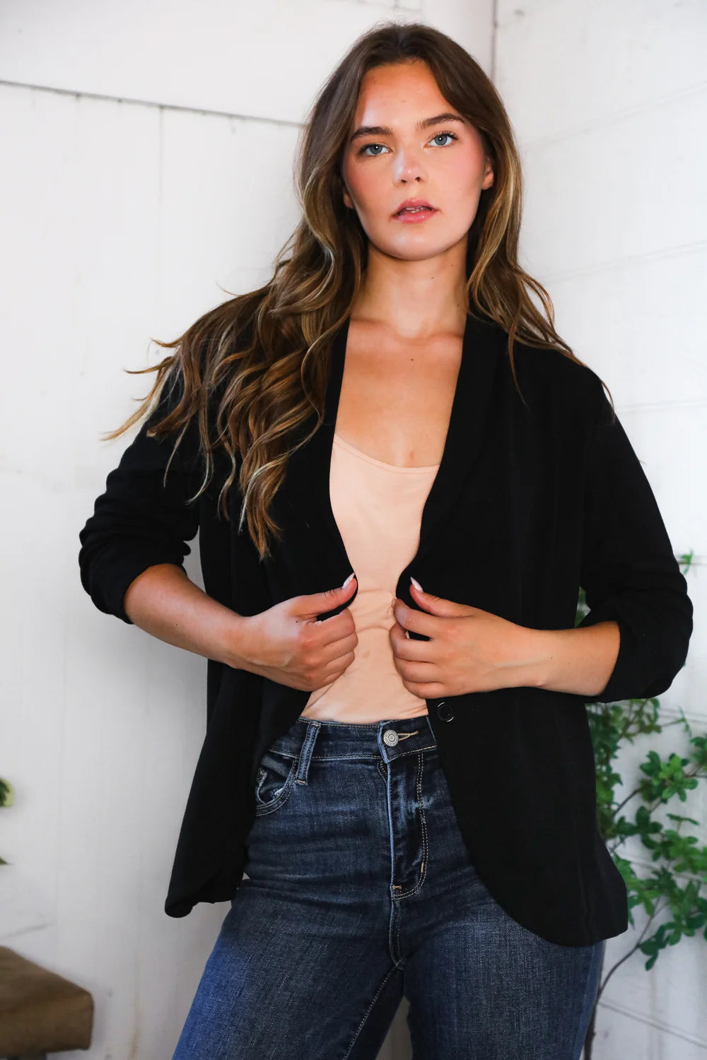 Woman wearing a black blazer over a beige top with blue jeans against a white wall.
