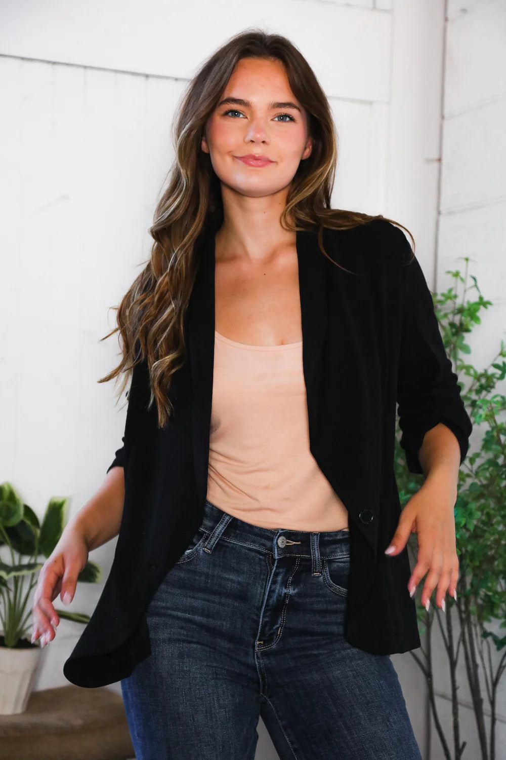 Woman wearing a black blazer over a beige top and blue jeans, standing indoors with plants in the background.