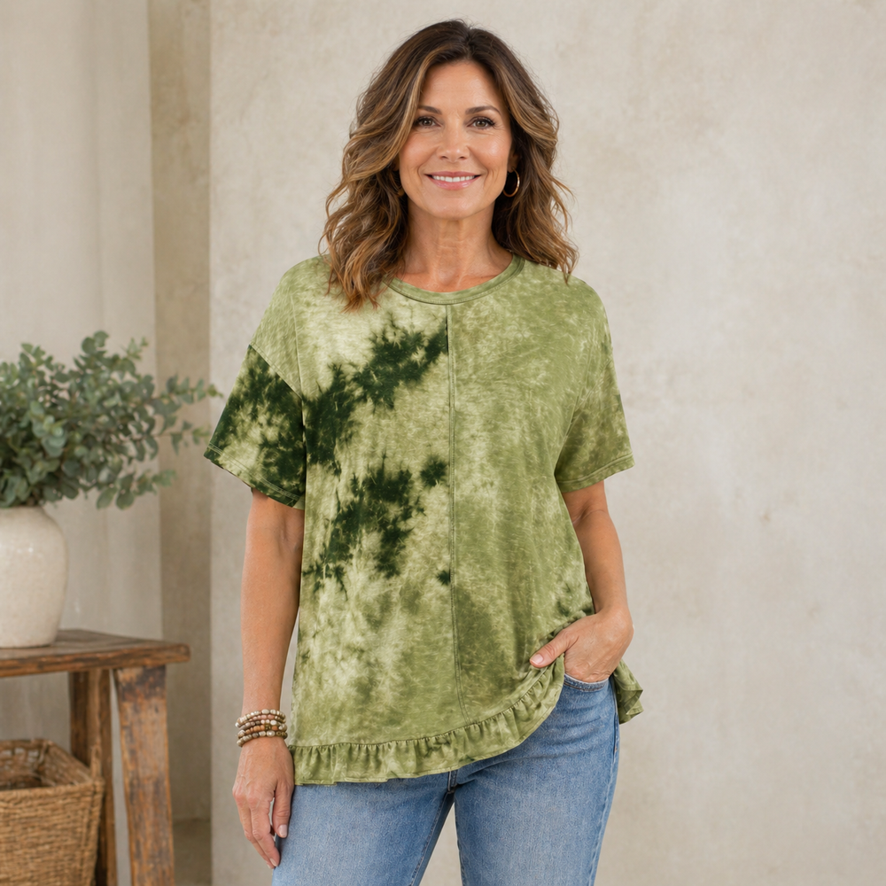 Woman wearing a green tie-dye shirt and blue jeans standing in a room with a plant and wooden table.