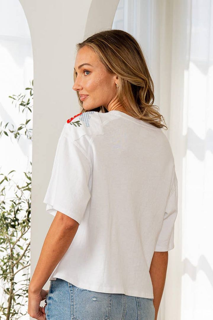 Woman wearing a white blouse with a red detail, standing outdoors with plants in the background.
