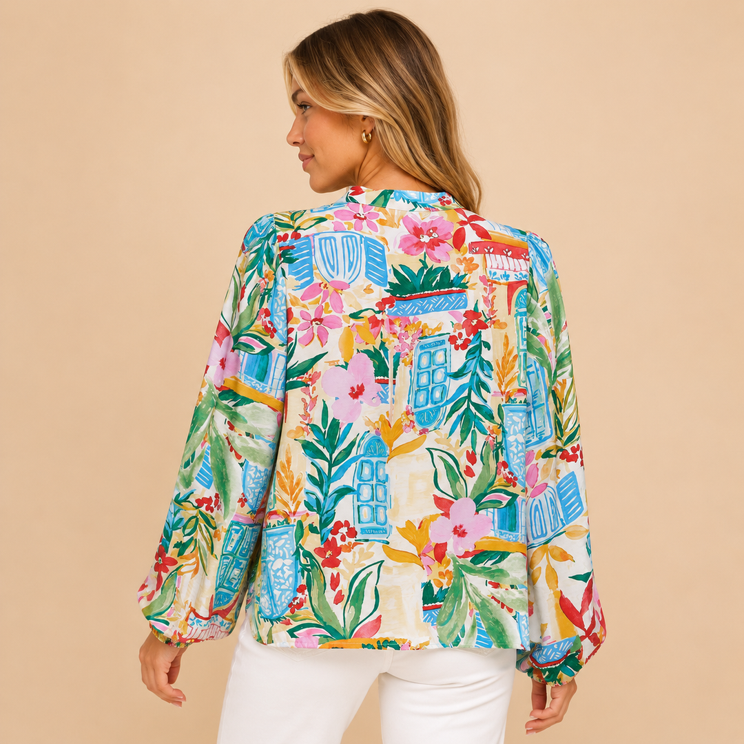 A woman shown from the back wearing a long-sleeve button-up blouse with a colorful tropical floral and architectural print, paired with white pants, against a solid beige background.