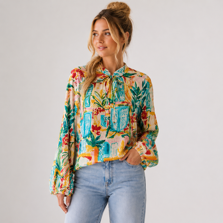 A woman wearing a long-sleeve tie-neck blouse with a colorful tropical and architectural print, paired with light-wash blue jeans, photographed against a plain light gray studio background.
