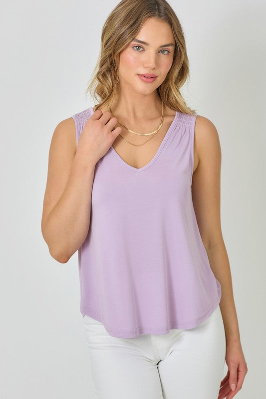 A woman wearing a lavendar sleeveless tank top with a v-neckline and smocked shoulders, paired with blue jeans.