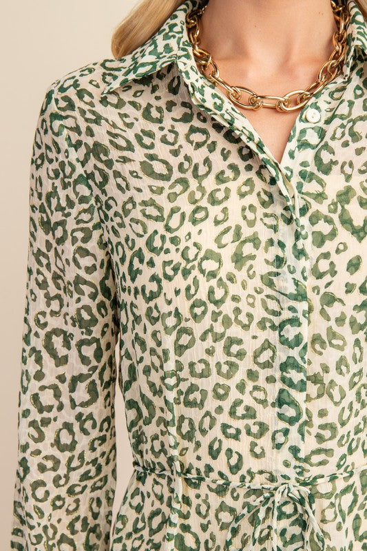A close-up of a woman wearing a long-sleeve collared shirt dress with a green leopard print on a cream background, styled with a chunky gold chain necklace.