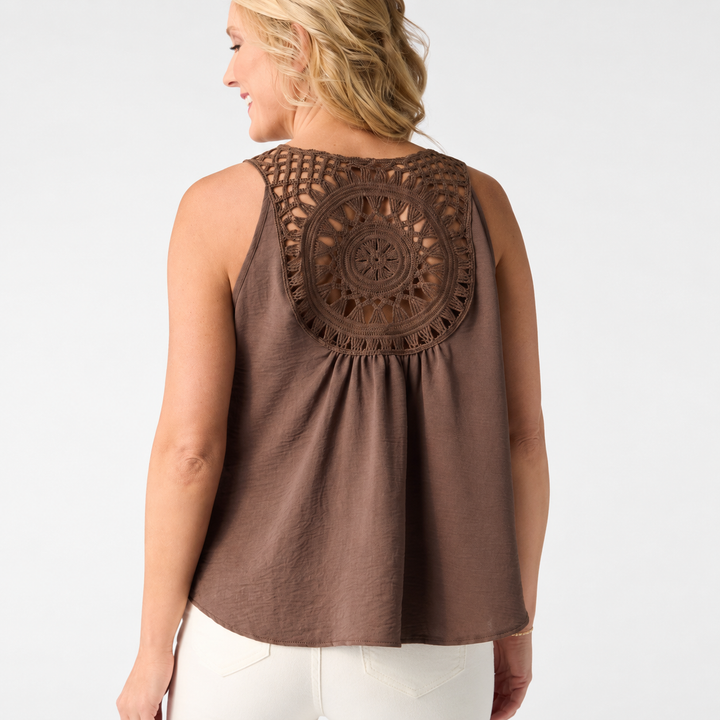 A woman shown from the back wearing a sleeveless brown top with a large circular crochet lace detail across the upper back, paired with white pants, against a plain light gray studio background.