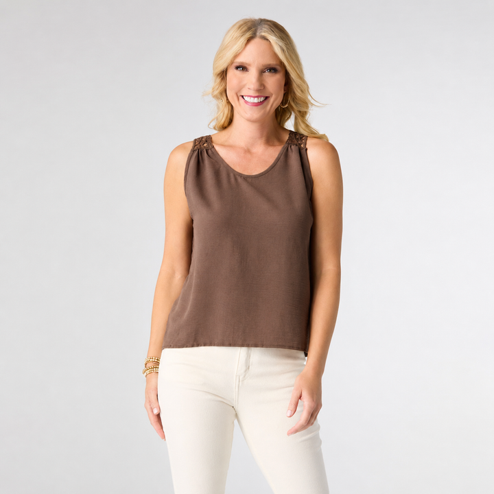 A smiling blonde woman wearing a sleeveless brown top with gathered shoulder details and white pants, standing against a light gray studio background.