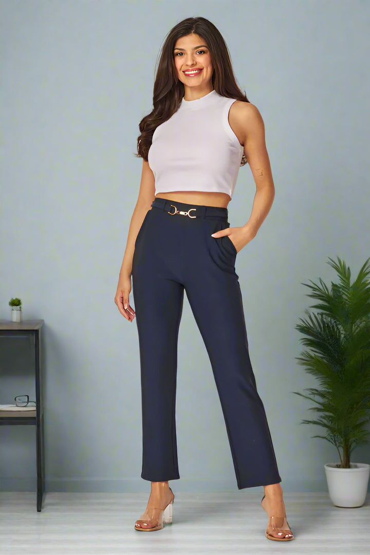 A model wearing tailored navy button cinched pants and a white sleeveless top, standing in a room with a potted plant and a furnished background.