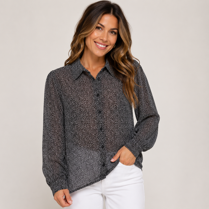 A woman wearing a sheer black and white speckled button-down blouse with white pants, standing against a light beige studio background.