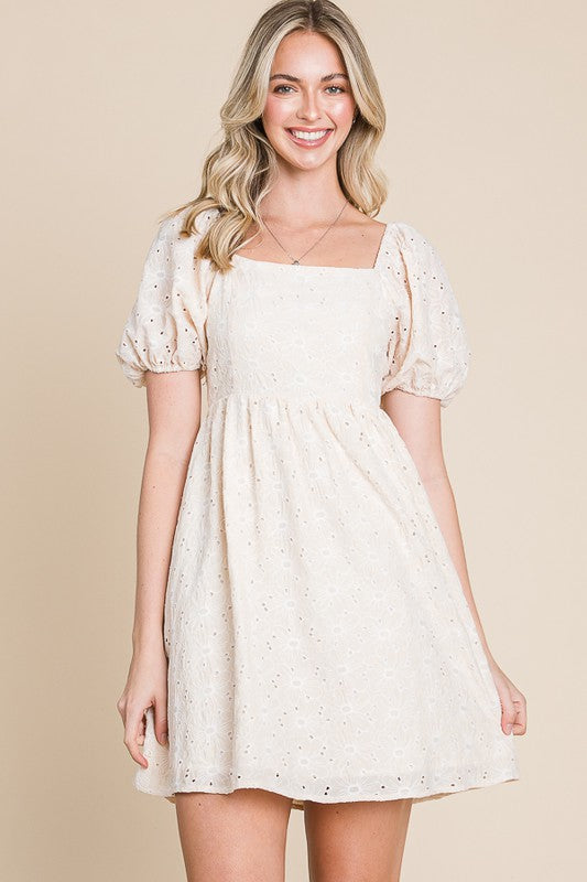 A woman wearing a beige mini dress with puff sleeves and eyelet detailing.
