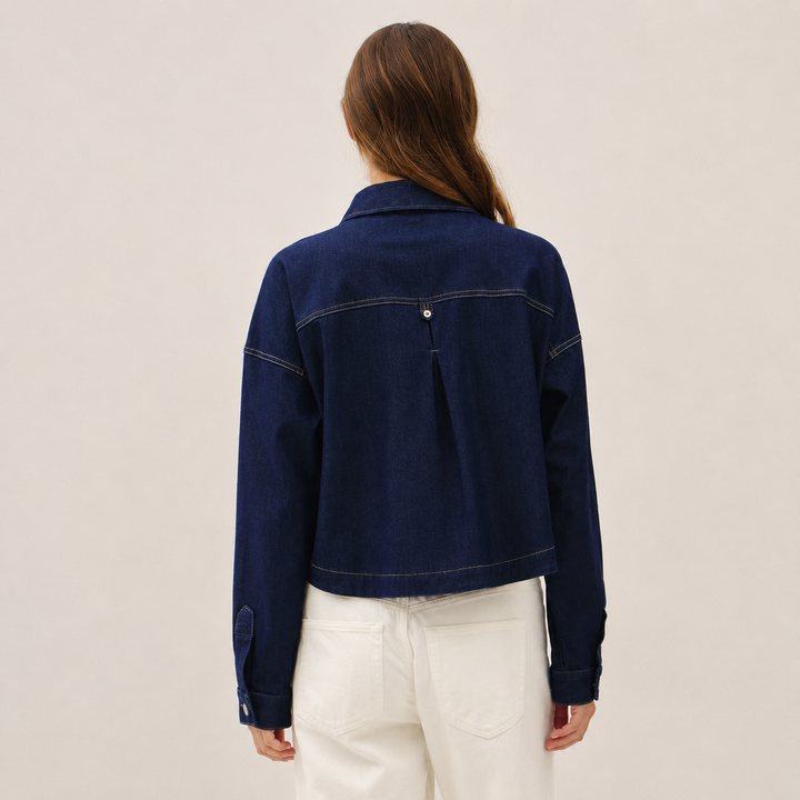 A woman viewed from the back wearing a cropped dark blue denim jacket with contrast stitching and white pants, set against a plain light beige studio background.