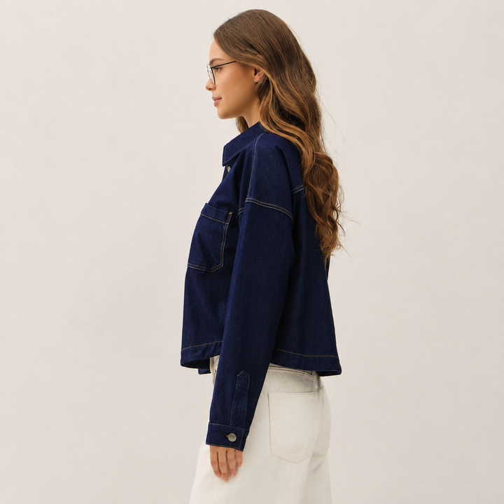 A woman in side profile wearing a dark blue cropped denim jacket with contrast stitching and white pants, set against a plain light beige studio background.