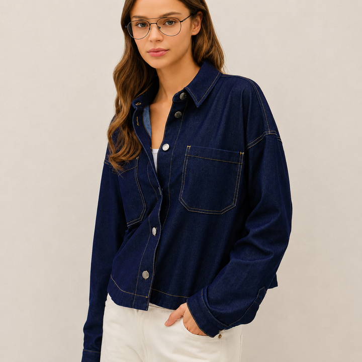 A woman wearing a dark navy blue denim button-up shirt jacket with white pants, photographed against a plain light beige studio background.