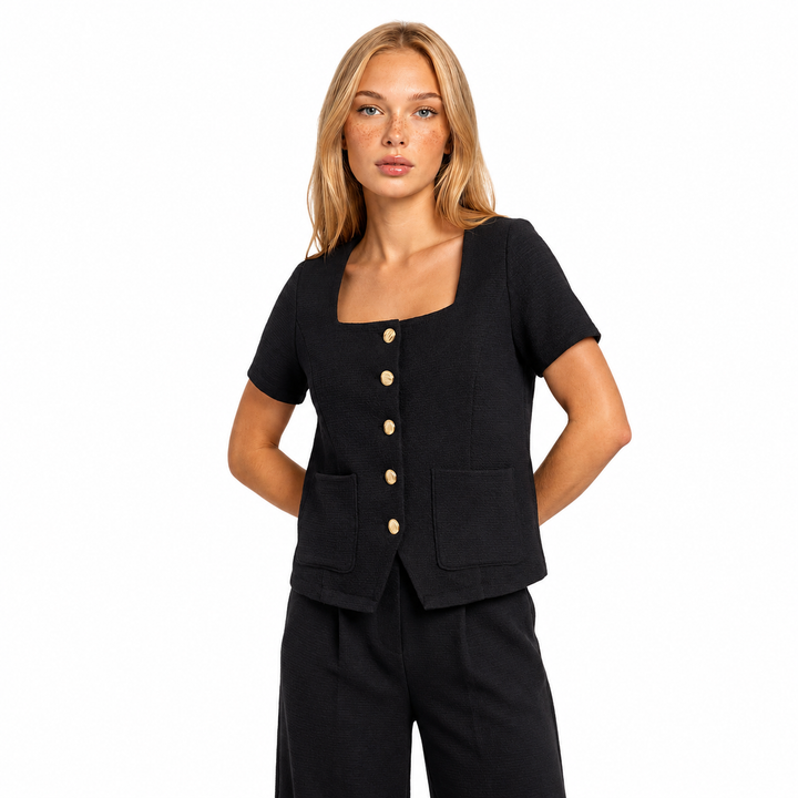 A woman modeling a black short-sleeve top with a square neckline, front button closure, and two front patch pockets, paired with black pants against a plain white background.