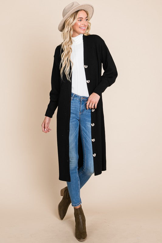 Woman wearing a black cardigan with button details, blue jeans, and brown boots on a beige background.