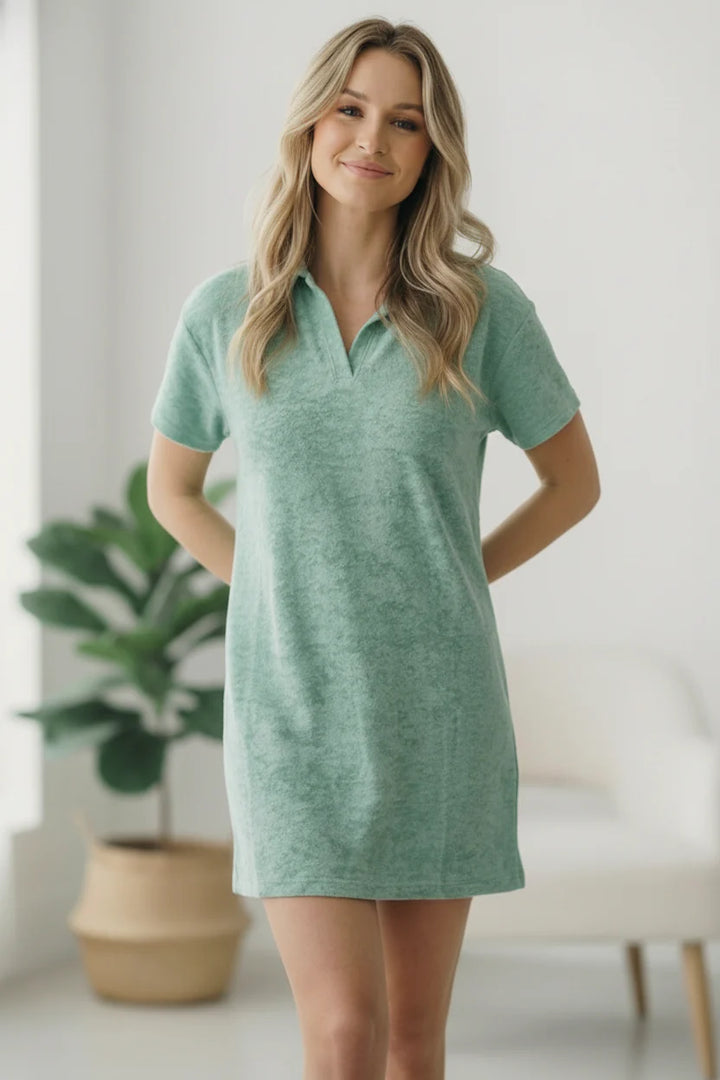 Woman wearing a green dress standing in a bright room with a plant in the background