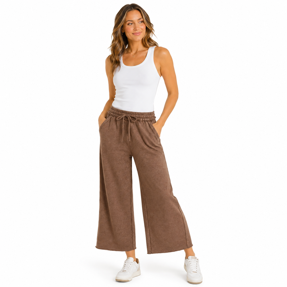 A woman modeling a pair of wide-leg brown pants with a drawstring waist, paired with a white tank top and white sneakers, against a solid white background.