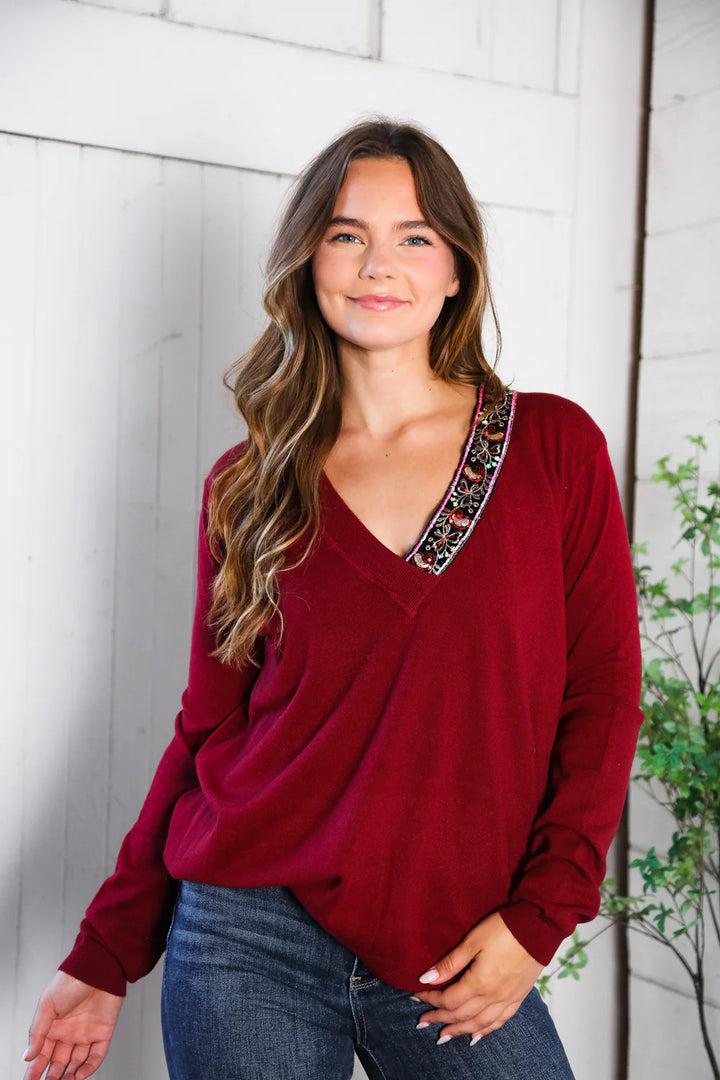 Woman wearing a wine long-sleeve top with a decorative neckline against a white brick wall.